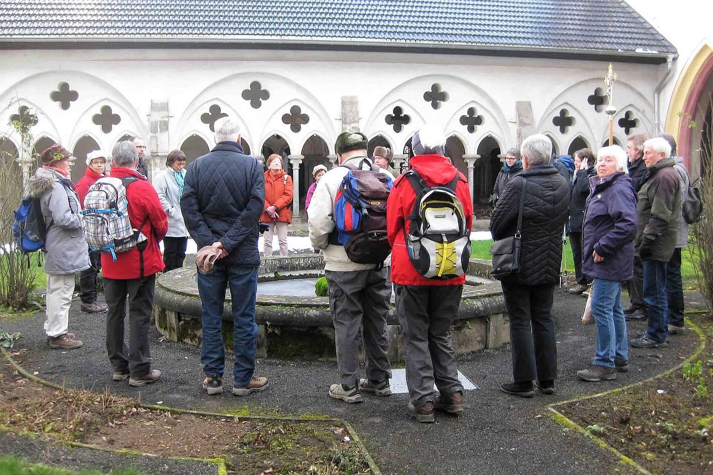 Ca. 20 Pilger*innen, z. T. mit geschulterten Rucksäcken und in warme Winterkleidung gehüllt, stehen rund um den Brunnen der Abtei Rommersdorf auf einem Schotterweg.