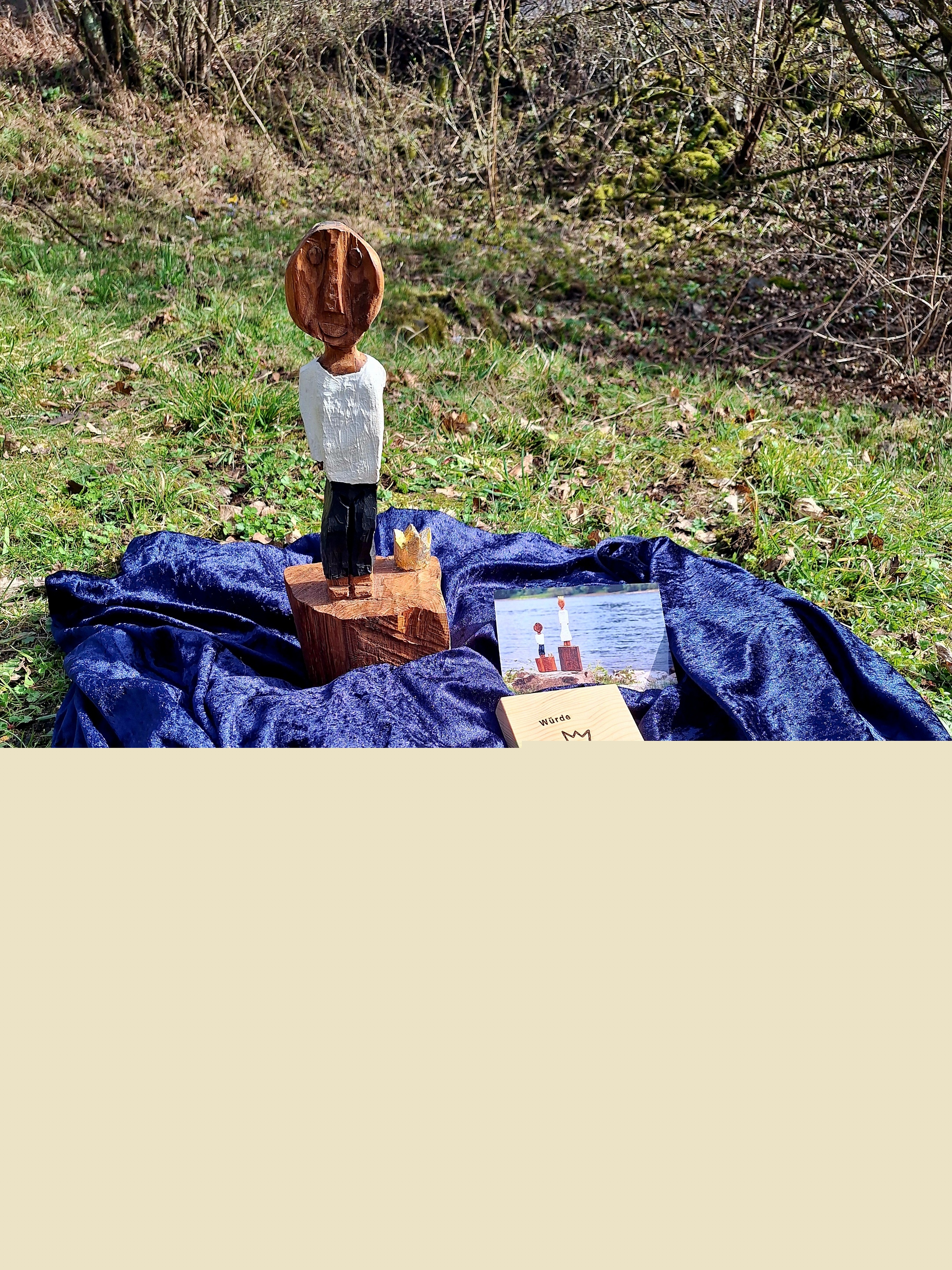 Auf einer Wiese steht die kleine Königsfigur auf einem blauen Pannesamt-Tuch. Auf dem Tuch liegen auch eine Würde-Tafel und eine Werbe-Postkarte.